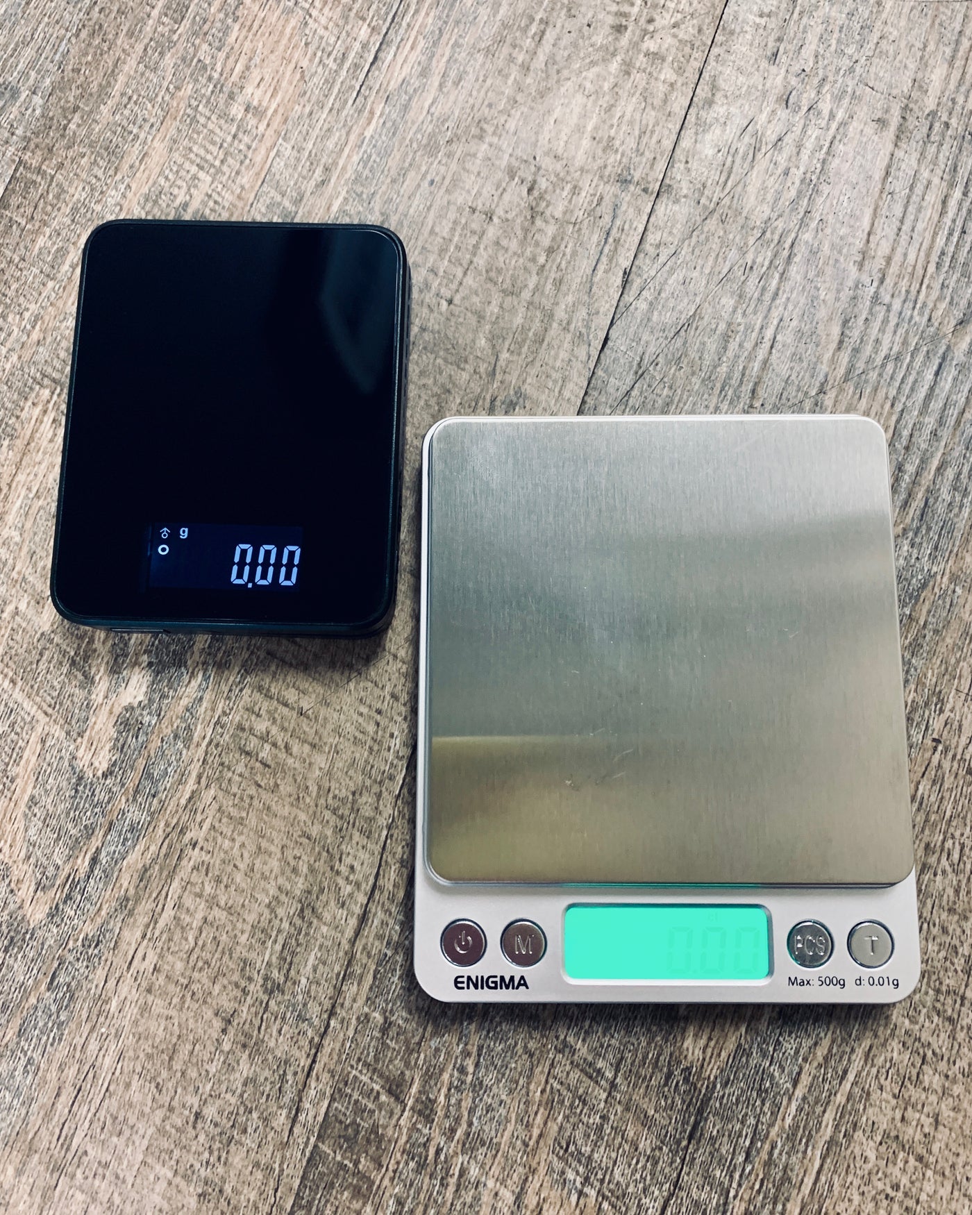 A Truweigh Storm and Truweigh Enigma sit on a wooden floor background. Both scales are turned on, the Storm is black with gray numbers and the Enigma has a green back light