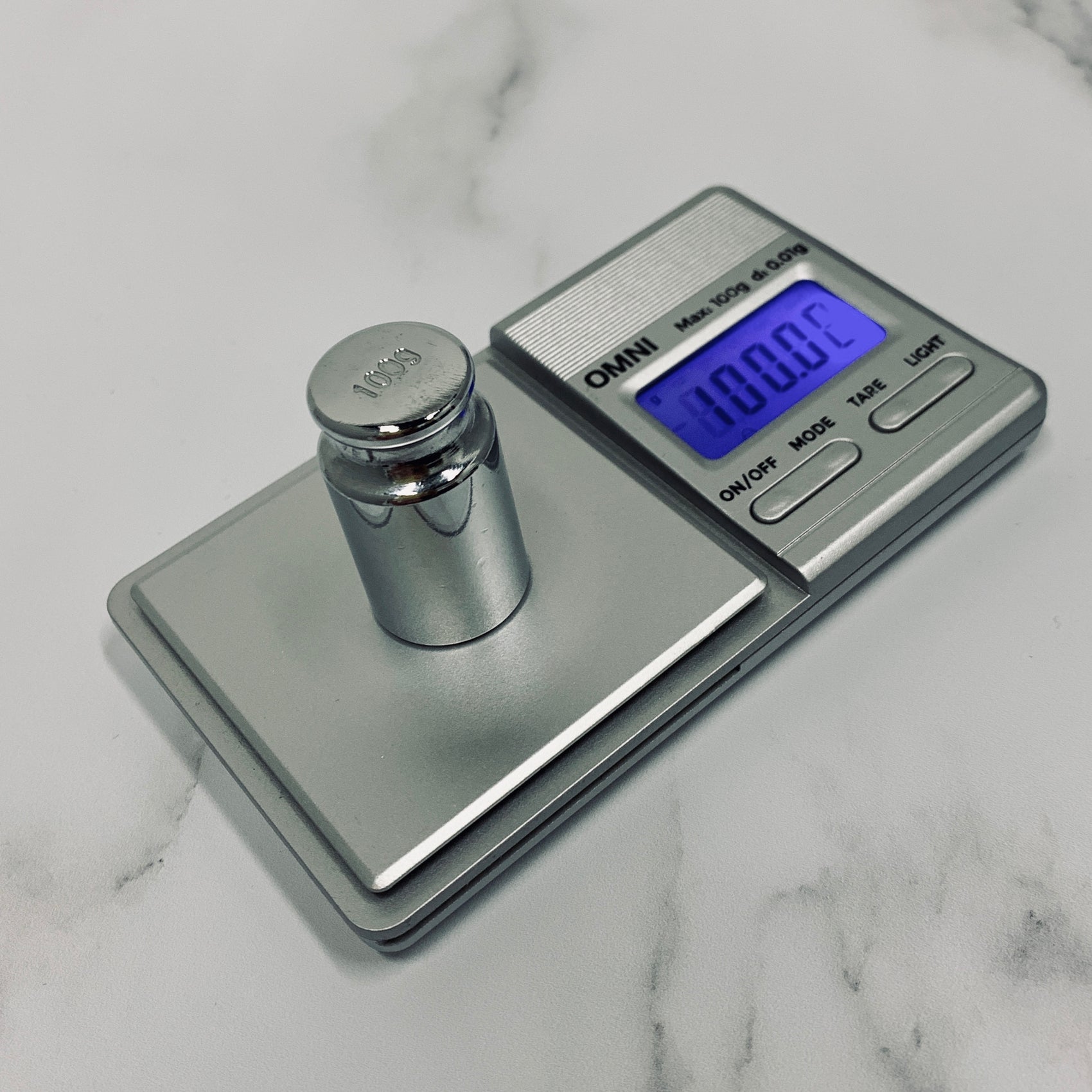 The Truweigh Omni Digital scale sits on a white marble background and is being calibrated with a 100g chrome calibration weight. The blue back-lit screen reads 100.00g.