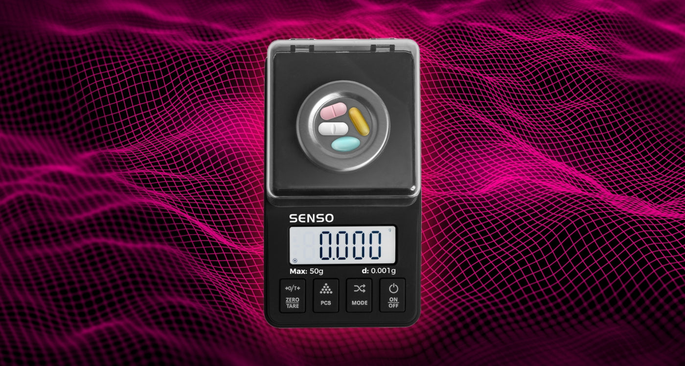 The Truweigh Echo Milligram digital scale sits on a marble counter, lid open with half of a pill on the weighing platform. The other half and a separate full pill sit on the stainless steel part of the scale.