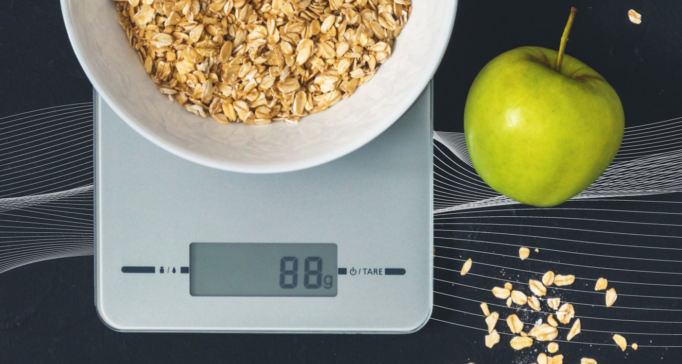 The Truweigh Crimson food scale sits on top of a pile of six meal prep containers that are filled with food. A large container of blueberries and a large container of strawberries are in front. Behind, a bunch of bananas and the Truweigh Vortex kitchen sc